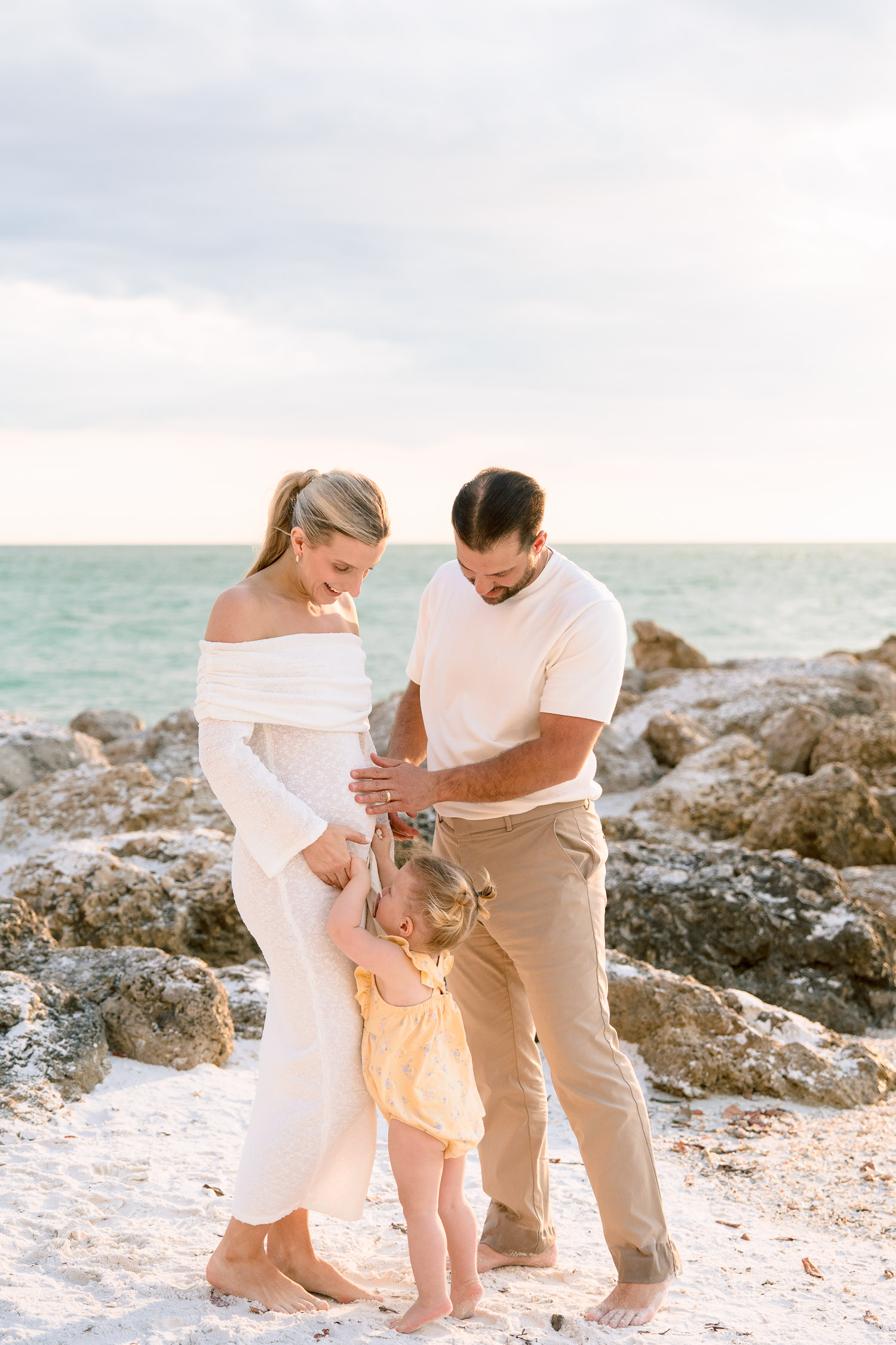 A glowing mother-to-be in white stands embraced by her family on the shore, framed by ocean waves and soft autumn clouds.