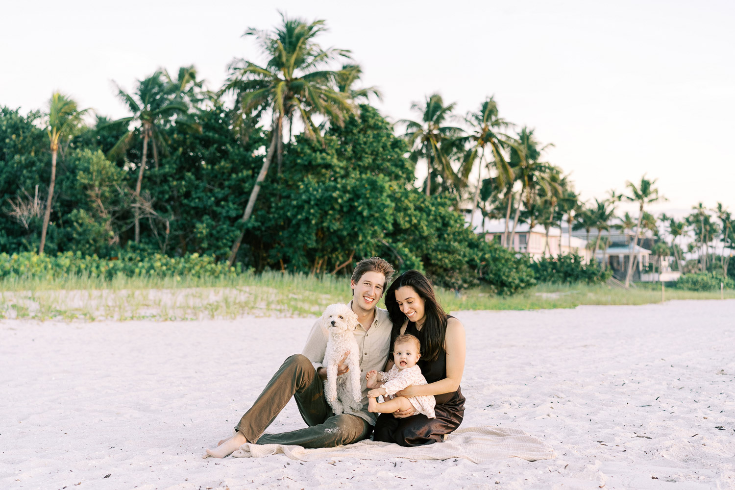 A joyful family with their baby and little dog share a sunlit embrace on the sand, framed by palms in coordinated autumn elegance.