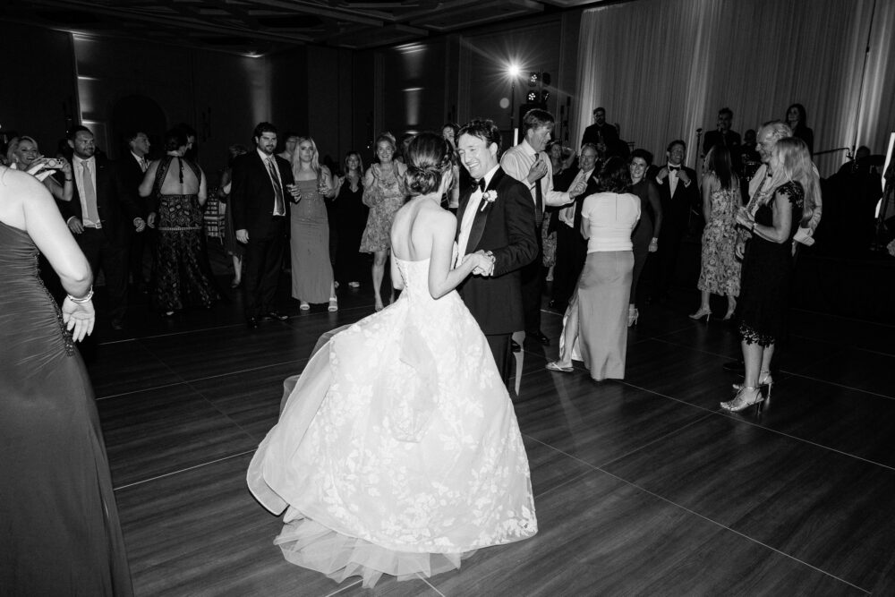The bride and groom share a romantic first dance as guests smile warmly, an elegant black and white moment in the grand ballroom at the Ritz Carlton Naples.